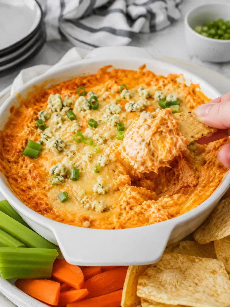 Close-up of Buffalo Chicken Dip in a white baking dish with melted cheese and blue cheese crumble, garnished with spring onion. A hand dips a crispy nacho into the creamy, spicy dip. Served with carrot, celery, and chips in natural light.